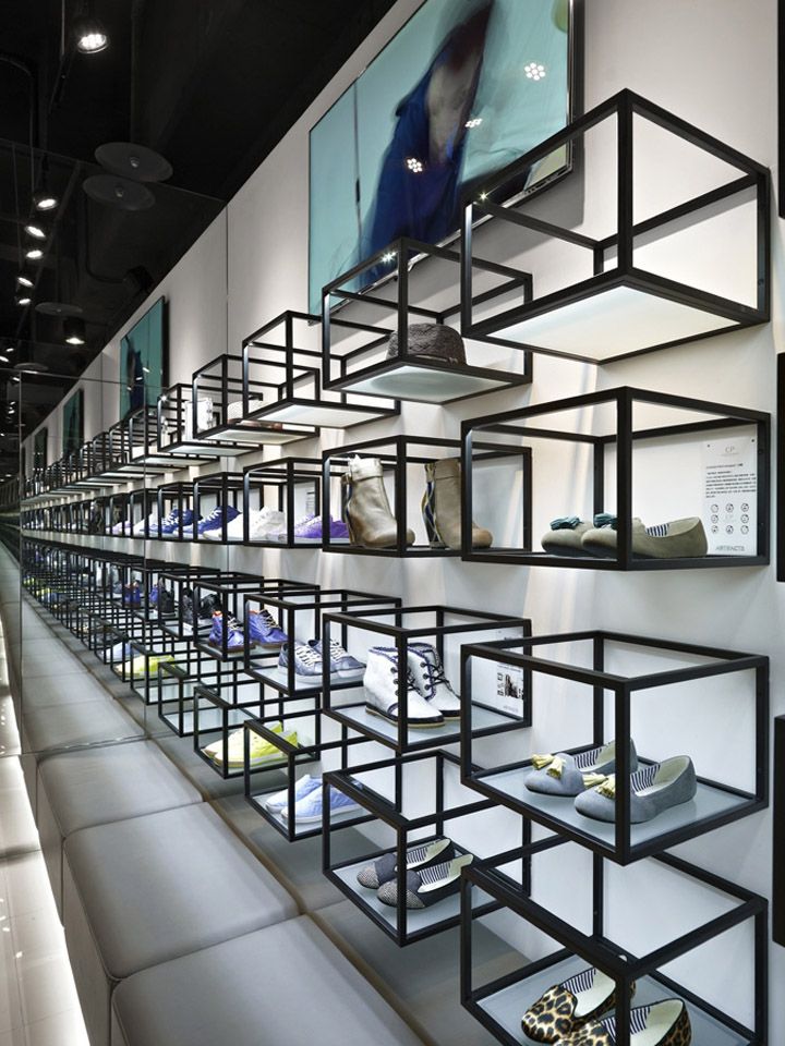 Black metal cube display shelves mounted on a white retail wall, holding various shoes at a footwear brand outlet, with a long grey bench below and spotlights overhead.